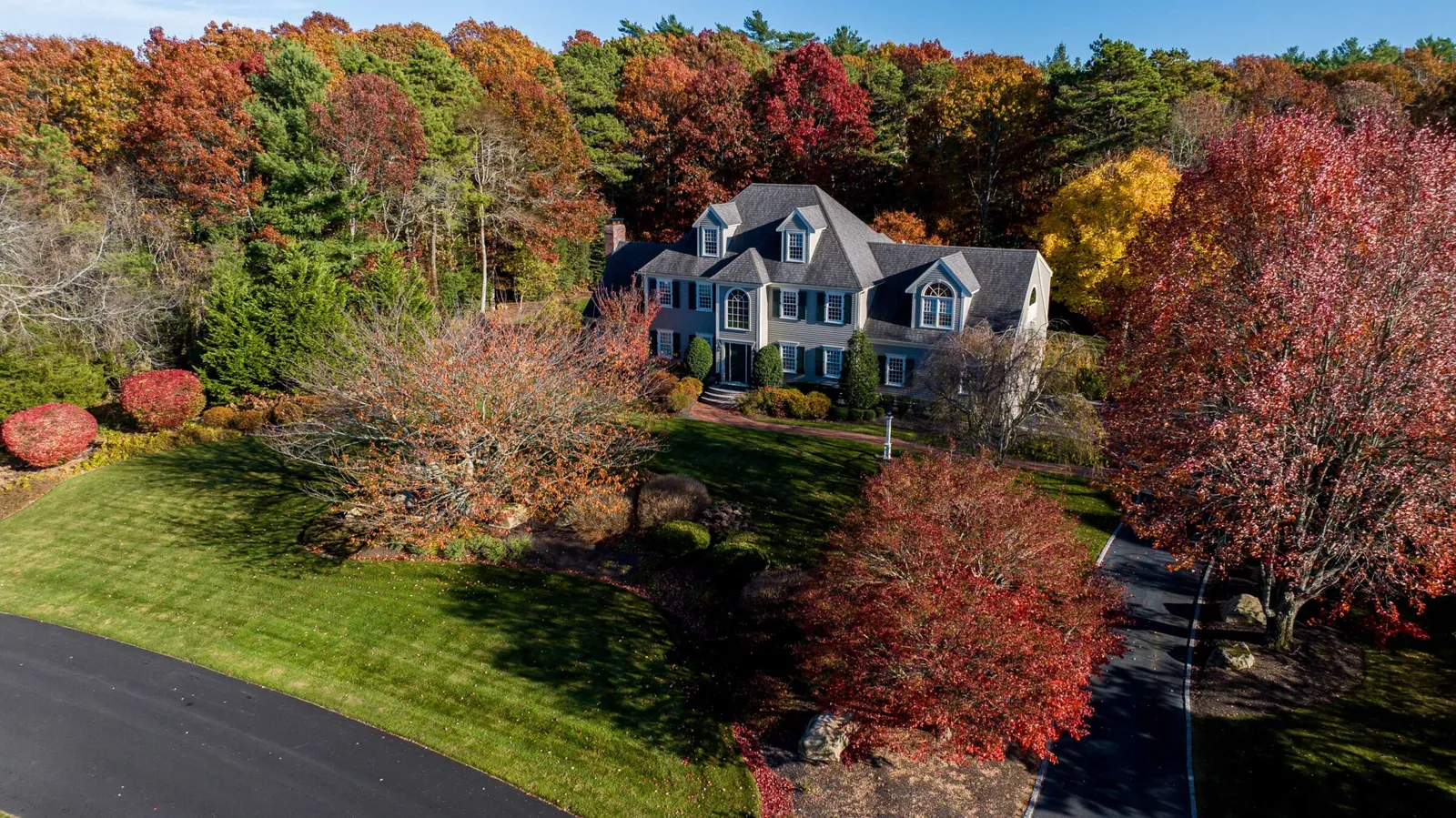 Aerial view of a large house surrounded by vibrant autumn trees, with a curved driveway and well-maintained lawn in a wooded setting.