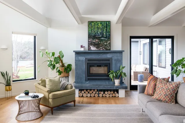 Modern living room with a fireplace, green armchair, indoor plants, coffee table, and large window overlooking a garden. Includes a gray sofa with patterned cushions and a painting above the mantel.