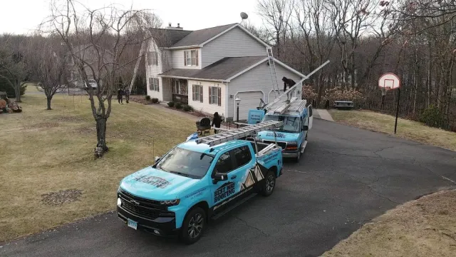 Blue truck with a ladder and aerial lift in front of a house. Men work on the roof.