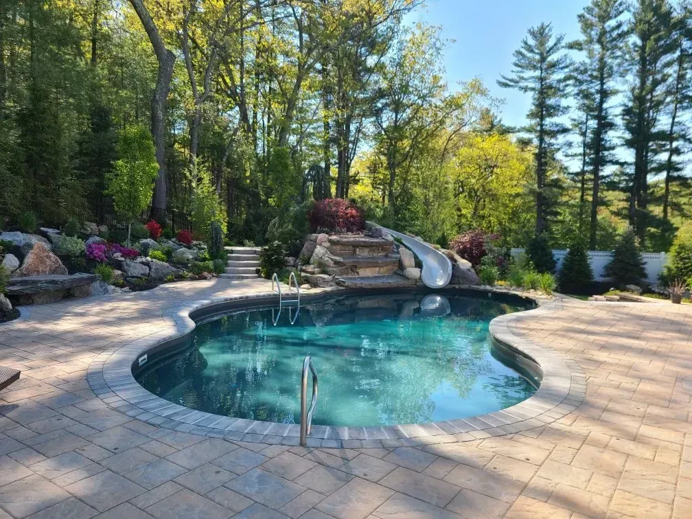 A kidney-shaped swimming pool with a stone slide and patio, surrounded by lush trees and a landscaped garden.