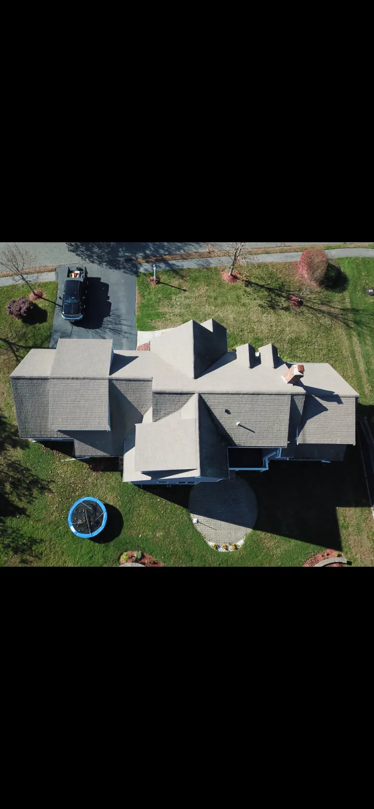Top-down aerial view of a house with a grey roof, a small trampoline in the backyard, and a car parked in the driveway, with trees and grass surrounding the property.