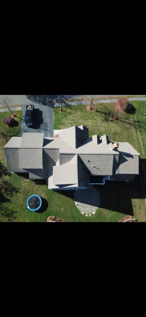 Top-down aerial view of a house with a grey roof, a small trampoline in the backyard, and a car parked in the driveway, with trees and grass surrounding the property.