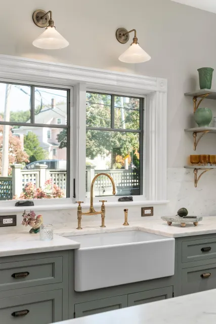 Bright kitchen with farmhouse sink, brass faucet, white countertops, and window overlooking greenery