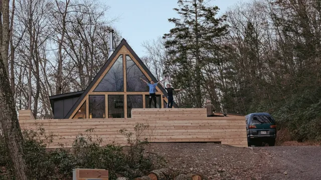 People outside on a wooden deck in front of a modern A-frame house, surrounded by trees, with a car parked nearby.