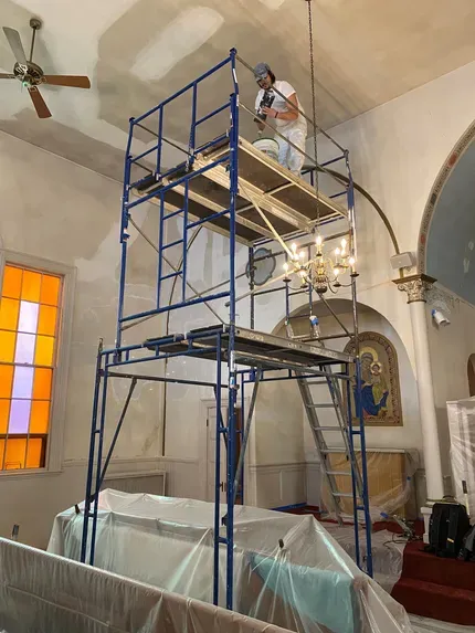 Worker on blue scaffolding inside a church, near stained glass and a chandelier.