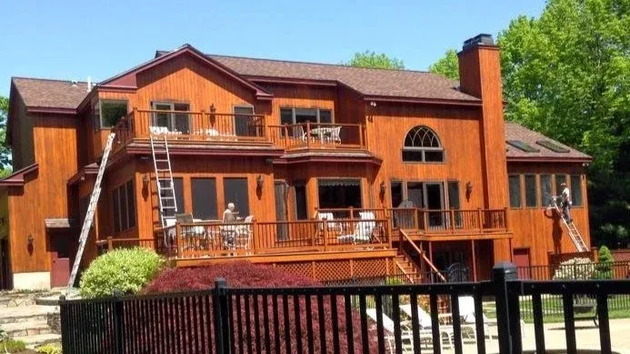 Large wooden house with multiple stories and a deck, surrounded by trees. Workers on ladders appear to be working on the exterior. Black fence in the foreground.