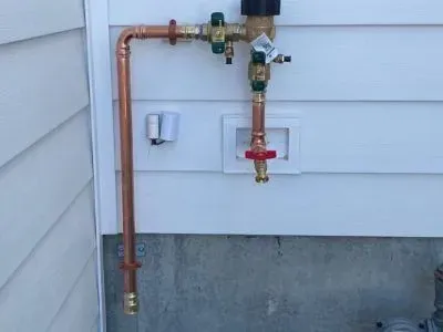 Copper piping on a white vinyl-sided wall, featuring a vacuum breaker valve and a water spigot in a recessed box.