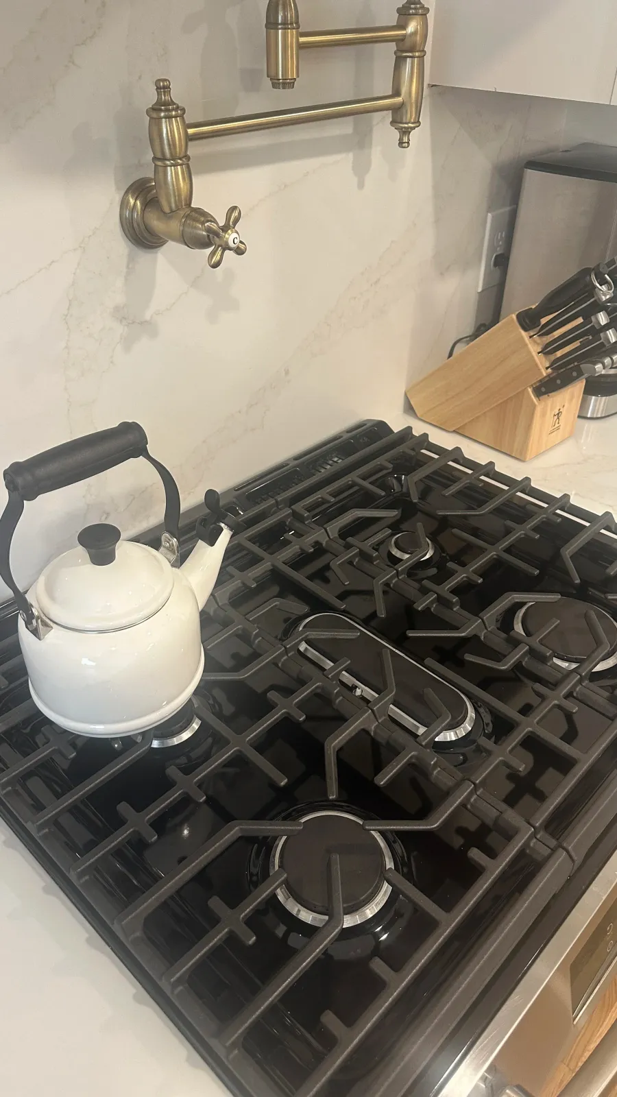 A close-up view of a kitchen stove with a white kettle placed on one of the burners, a knife block with knives in the background, and a brass faucet mounted on the wall.