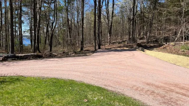 Gravel driveway curving through a wooded area with grass in the foreground and water visible through trees