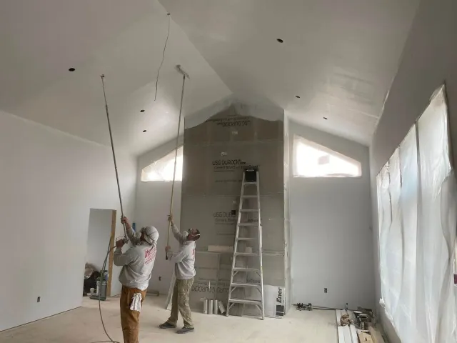 Two workers installing ceiling fixtures in a bright room with a ladder and plastic-covered wall.