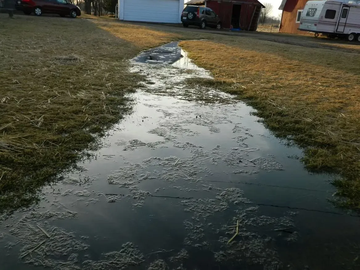 A long, shallow, water-filled ditch cuts through a grassy yard. Buildings and vehicles are in the background.
