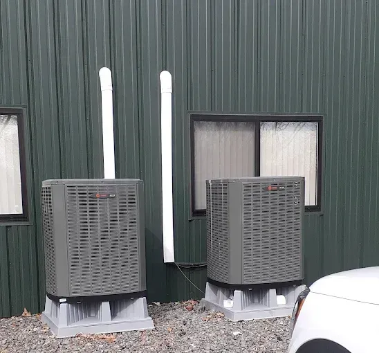Two gray HVAC units with white vent pipes outside a green building, next to a car.