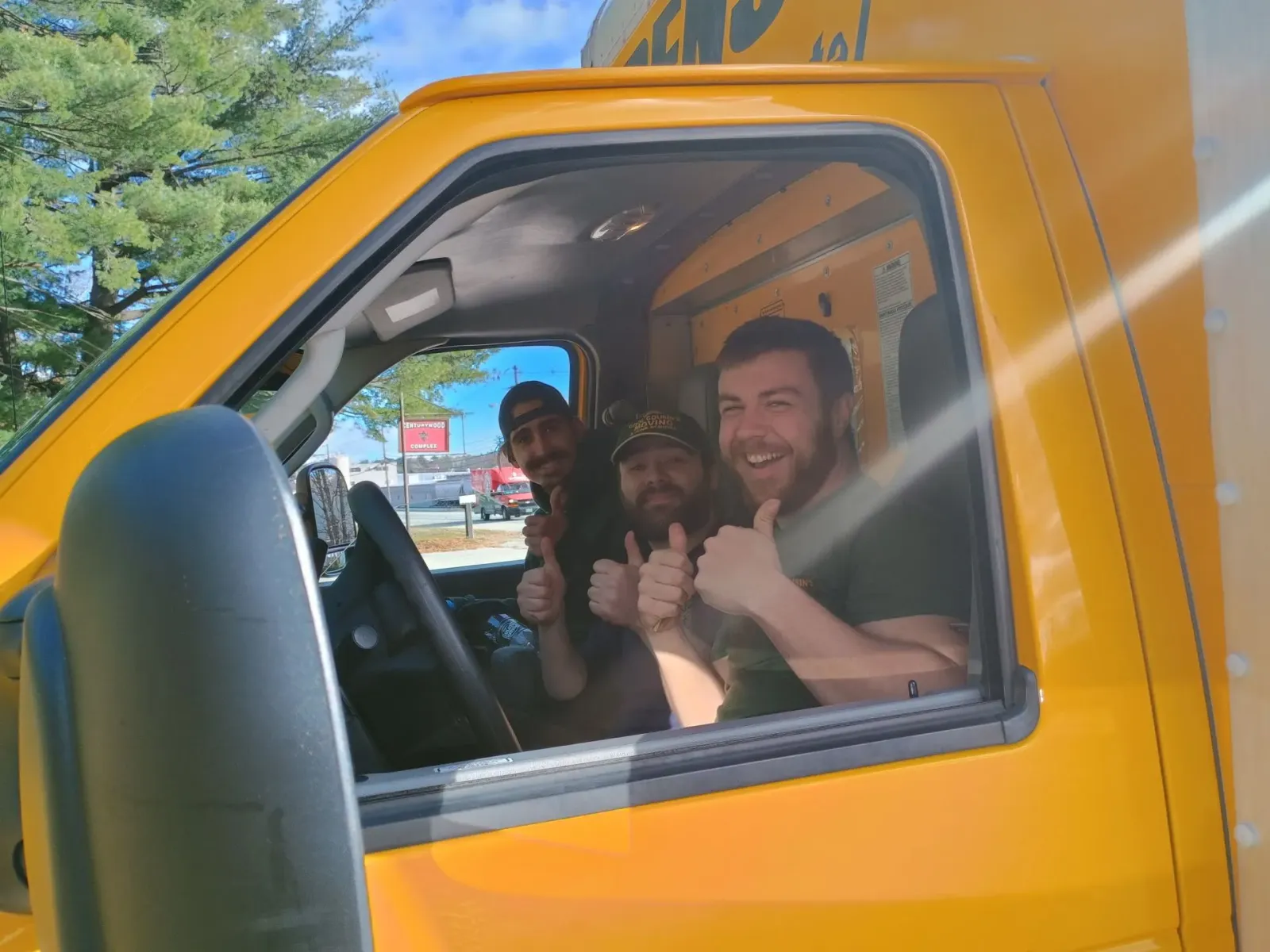 Three smiling people in an orange van, one giving a thumbs-up through the window.