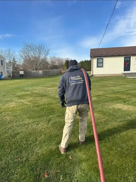 A person in a gray hooded sweatshirt and tan pants walks across a grassy yard while holding a long, red service hose.