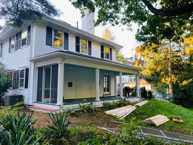 White two-story house with porch. Boards and tools on the grass, possibly under renovation.