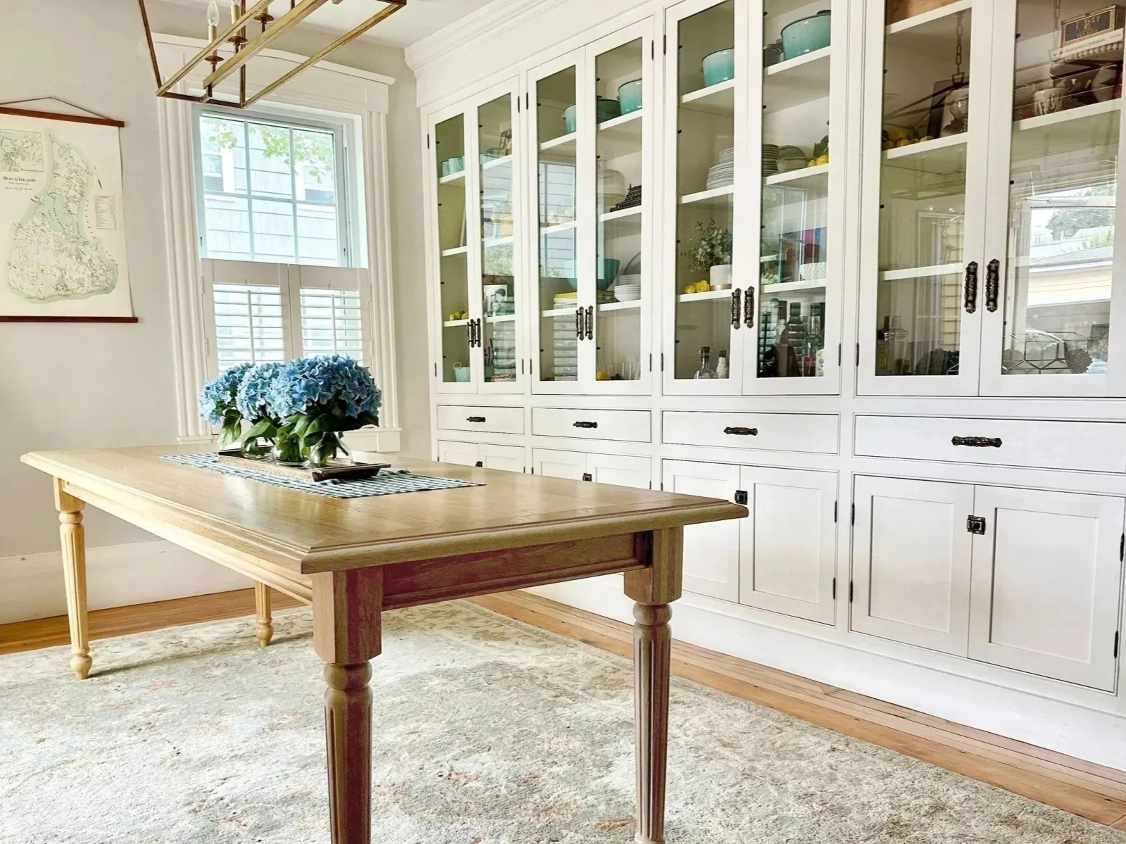 A dining area with a wooden table, a glass-fronted white cabinet filled with dishes and decorative items, a window with shutters, and a floral arrangement of blue hydrangeas on the table.