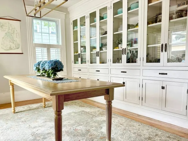 A dining area with a wooden table, a glass-fronted white cabinet filled with dishes and decorative items, a window with shutters, and a floral arrangement of blue hydrangeas on the table.