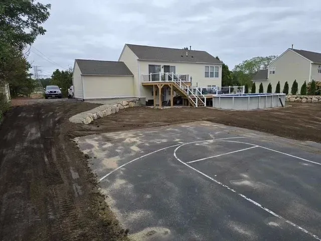 House with deck and basketball court beside a muddy driveway, viewed from a dirt road.