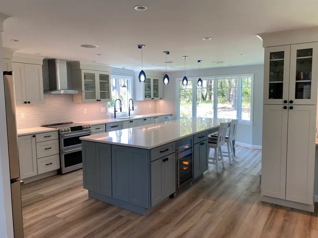 Bright, modern kitchen with white cabinets, a center island with a wine fridge, stainless steel stove, and large windows overlooking a wooded yard.