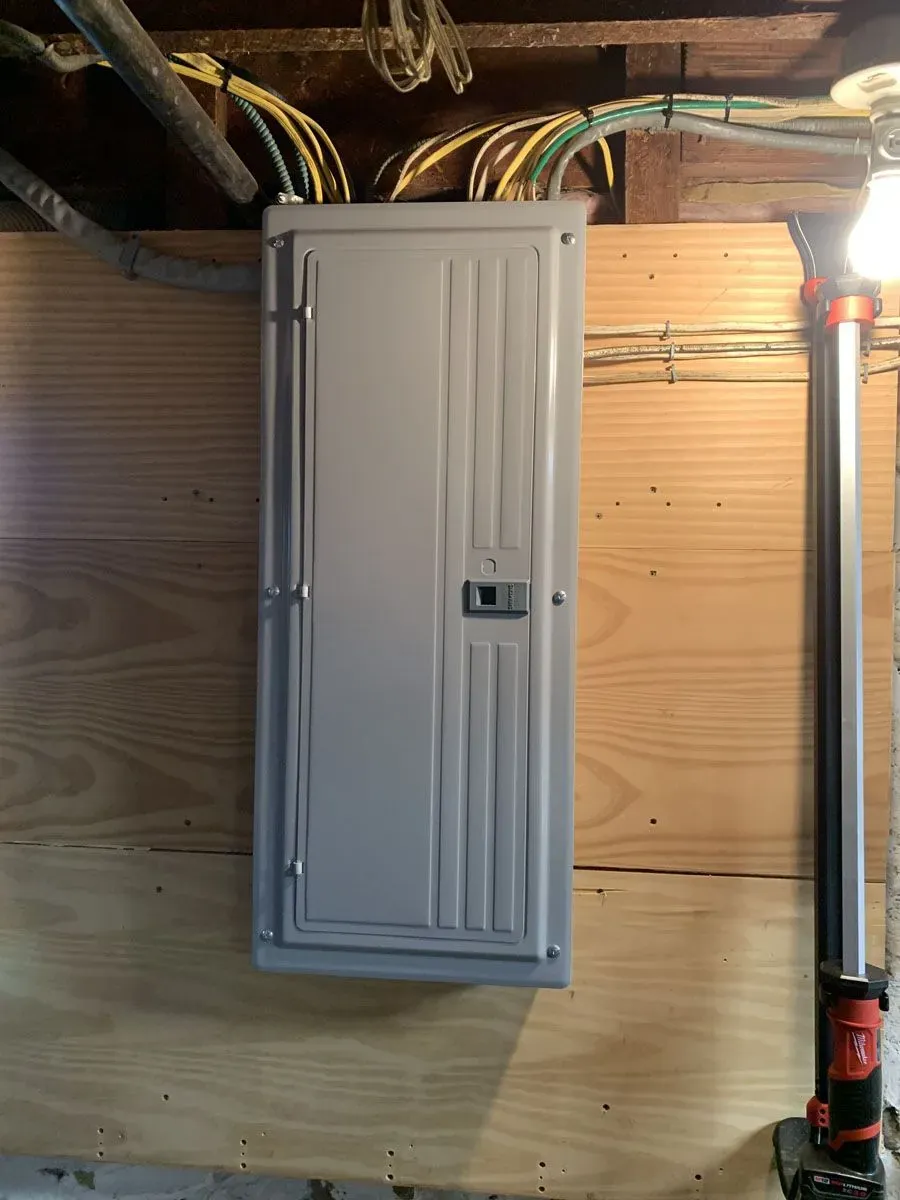 A gray electrical panel mounted on a plywood wall in a basement. Wiring and a work light are also present.