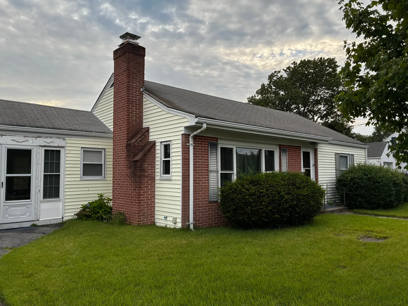 A house with a brick chimney, yellow siding, white trim, and a slate roof, with a grassy lawn and trees in the background under a cloudy sky.