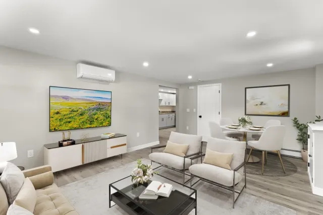 Modern living room with white walls, beige and white furniture, a wall-mounted TV, and a dining area with a round table and four chairs.