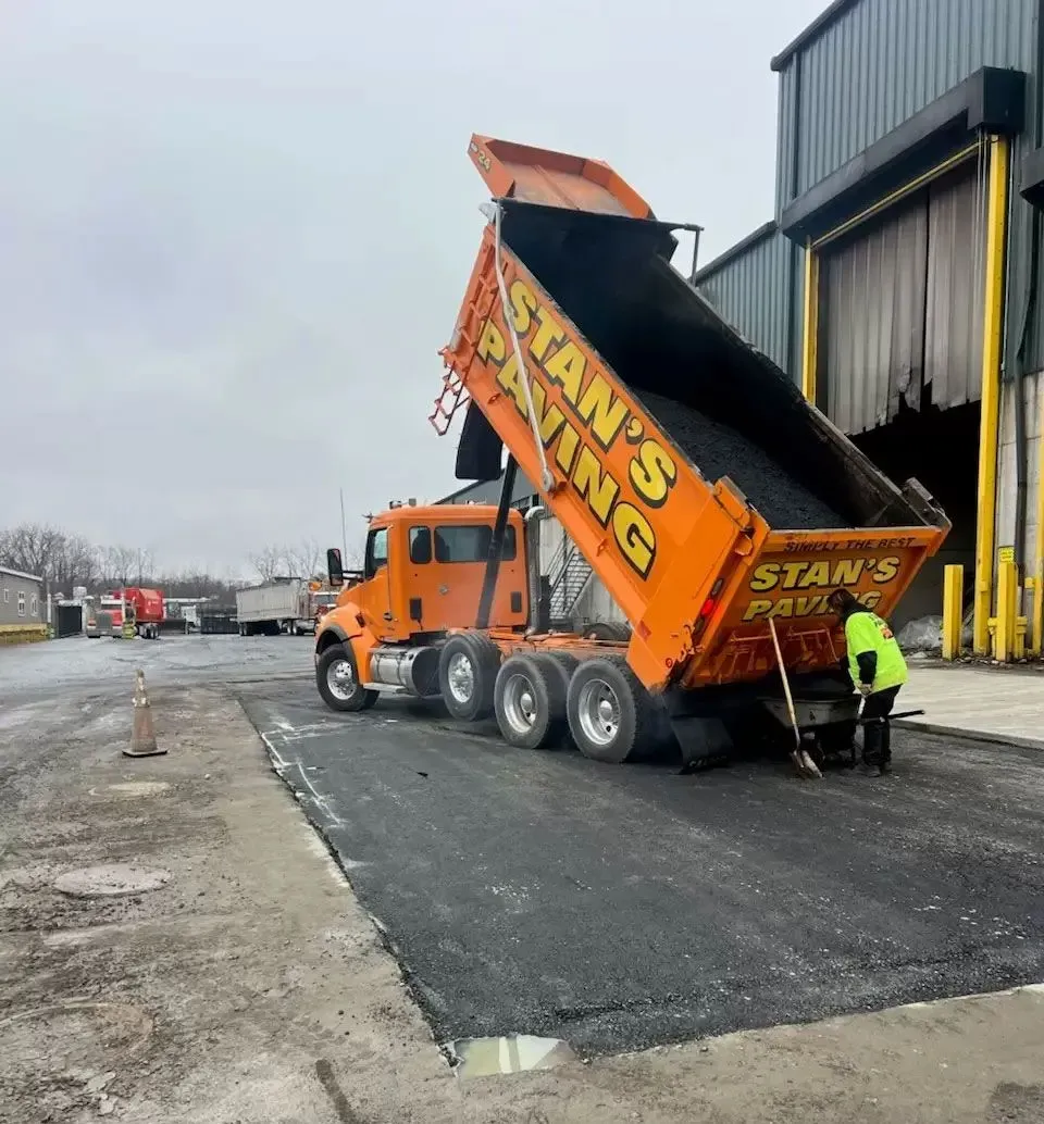 Orange dump truck tipped up at a construction site, unloading material near a warehouse loading dock.