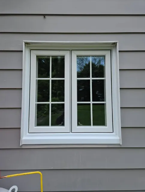White-framed double window on a gray clapboard exterior, reflecting trees and sky.
