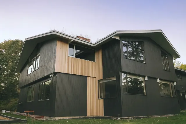 A modern two-story house with black and natural wood siding, large windows, and a sloped roof surrounded by trees.