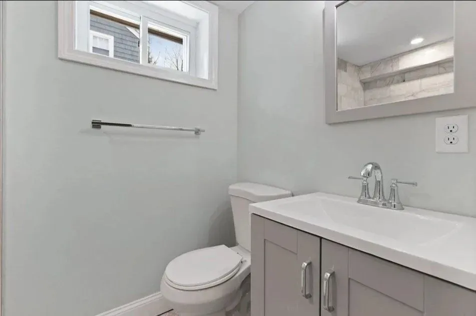 A bathroom with a toilet, sink, mirror and window.
