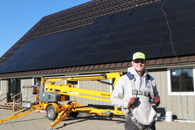 A man wearing a green cap, gray hoodie, and safety harness working on installing solar panels on a building's roof with a lift in the background.