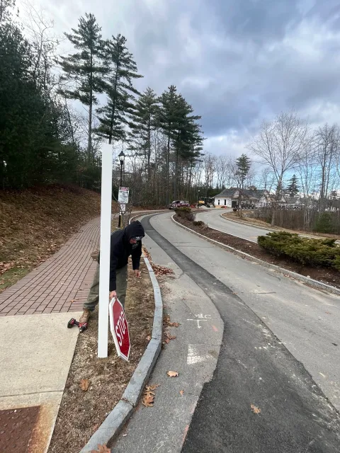 A person in a black jacket and beige pants is bending down on a sidewalk, holding a stop sign. There is an upside-down stop sign leaning against a white pole nearby. The scene is outdoors in a neighborhood with trees, houses, parked cars, and a cloud