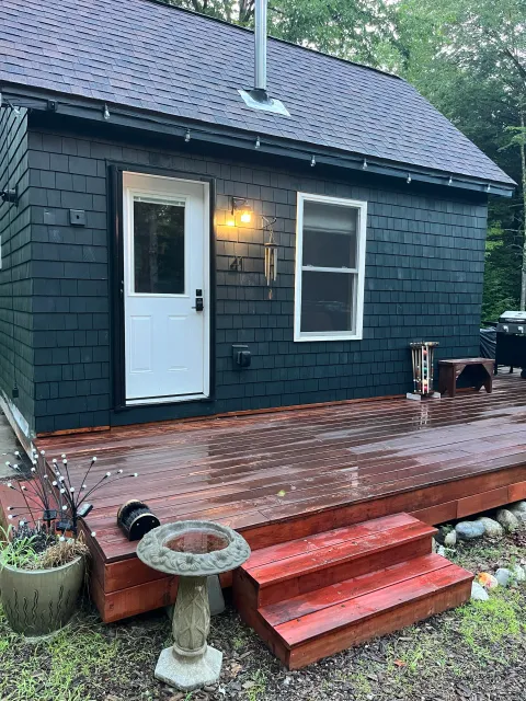 A wooden deck attached to a house with black shingles exterior siding, two windows, a door with a keypad lock, and outdoor decorations including a lamp, wind chimes, a small bench, and potted plants. The deck has steps leading down to a grassy yard.