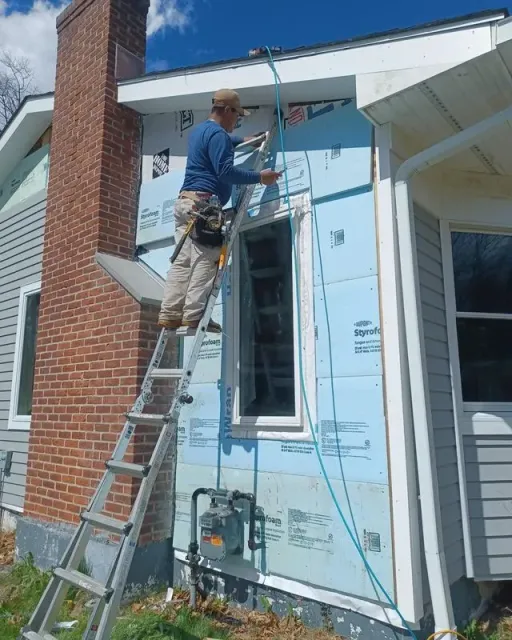 Person on ladder installing siding on house exterior, blue insulation visible.