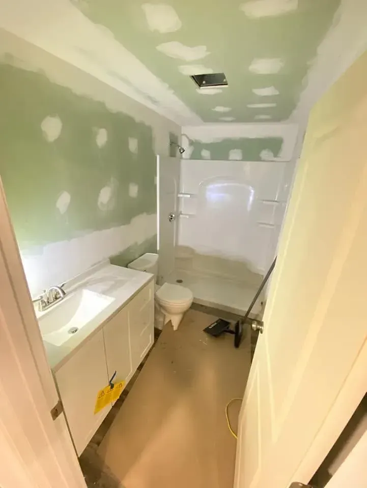 Narrow bathroom with green drywall, white sink vanity, toilet, bathtub, and unfinished ceiling patching.