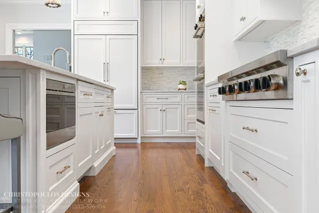White kitchen with wooden floors, white cabinets, stainless steel appliances, a small plant, and a window showing trees outside.