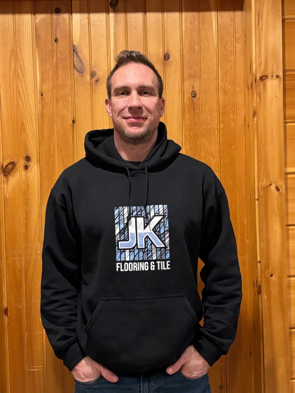 Business owner black hoodie with "JK Flooring & Tile" logo standing in front of wood panel wall.