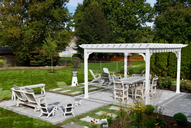 A backyard patio with a white pergola, outdoor furniture including chairs, benches, a dining table, and small side tables. There is a grassy lawn with a small tree, a garden area with plants and flowers, and a decorative lighthouse. The background fe