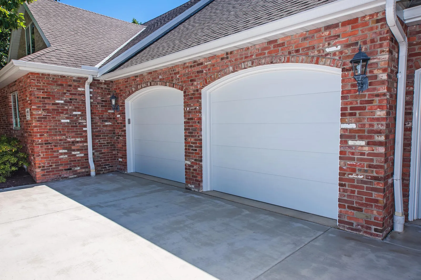 Brick house with two arched white garage doors under a gray shingled roof; concrete driveway in foreground.