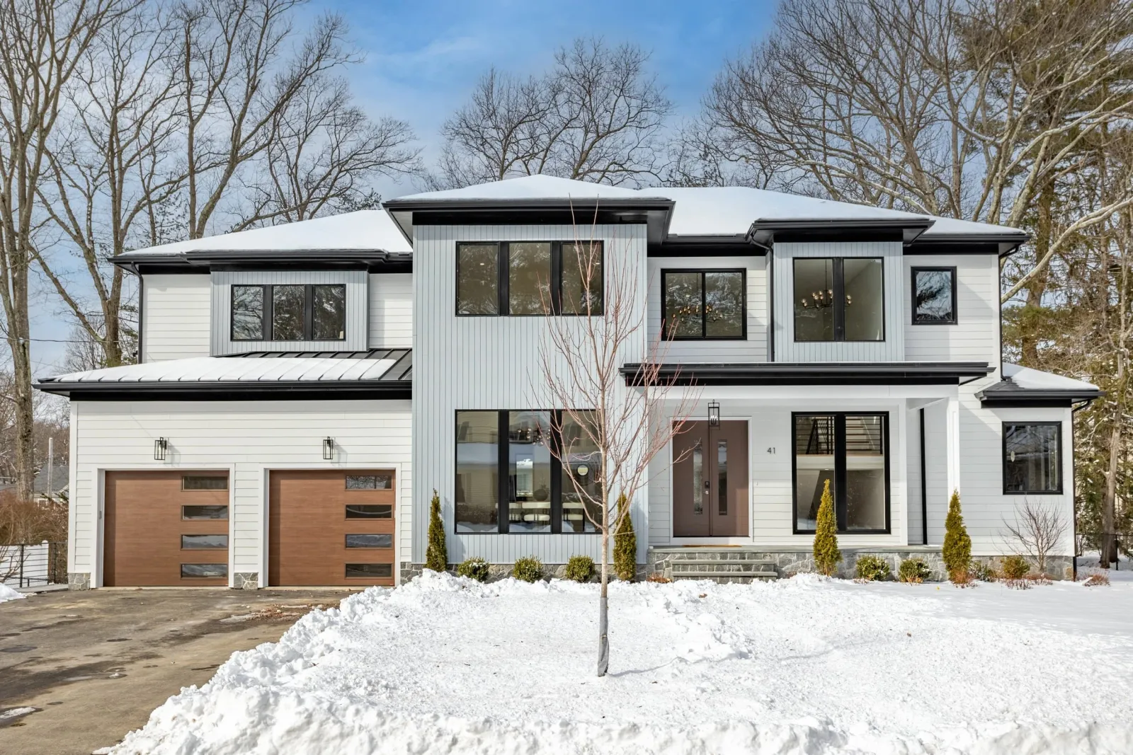 A modern two-story house with white and gray exterior, large windows, and a snow-covered yard. Features two brown garage doors and a single front door, with leafless trees in the background.