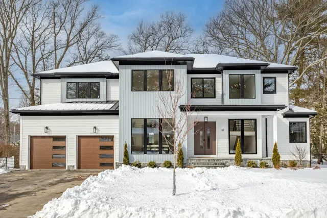 A modern two-story house with white and gray exterior, large windows, and a snow-covered yard. Features two brown garage doors and a single front door, with leafless trees in the background.