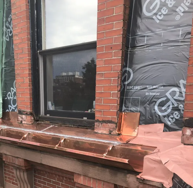 A brick building under construction with a black window and copper flashing around the window and on the roof ledge, with construction materials visible inside the window.. Work done by WP Walsh Roofing.