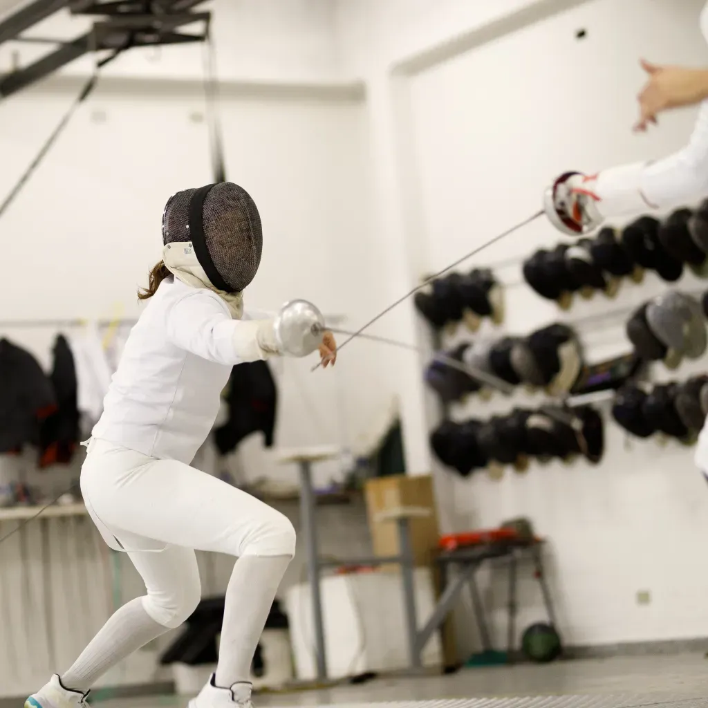 Female épée fencer performing a deep lunge during a bout at Allez Fencing Club in Watertown, MA, with fencing masks and gear visible in the background.