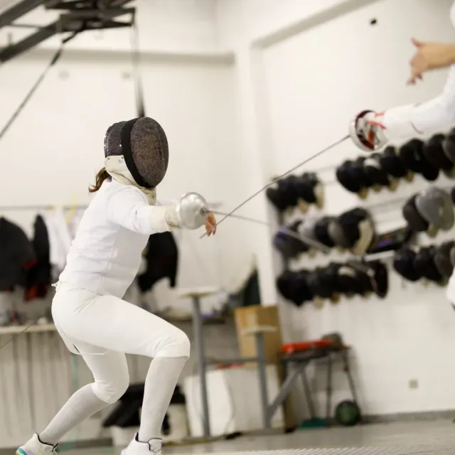 Female épée fencer performing a deep lunge during a bout at Allez Fencing Club in Watertown, MA, with fencing masks and gear visible in the background.