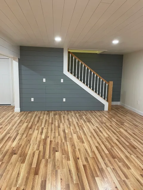 Empty living room with hardwood floors, a gray accent wall, and a staircase with white spindles and wooden handrail leading upstairs.