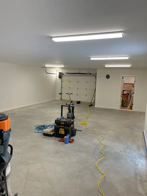 Empty garage with fluorescent lighting, open garage door, tools and equipment on the concrete floor, yellow extension cord, and a doorway leading into another room.