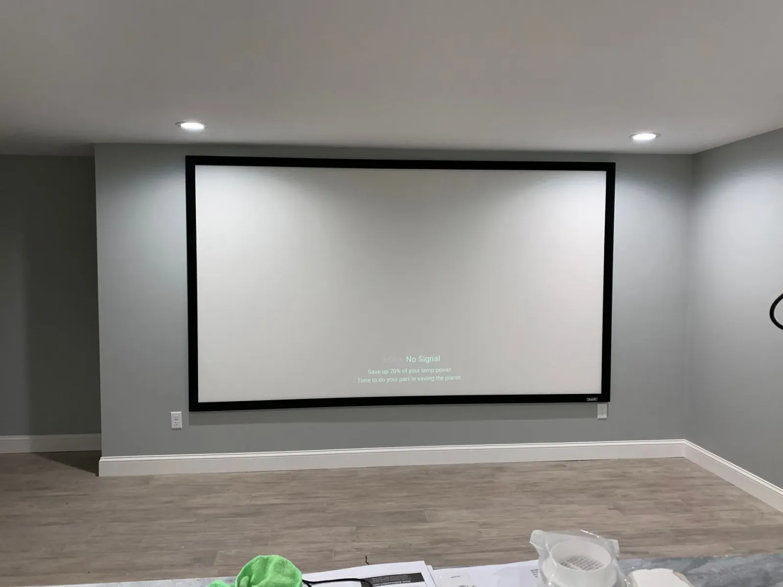 A large rectangular projector screen with a black frame mounted on a light gray wall in a room with wood-look flooring.