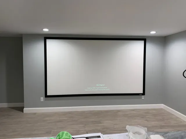A large rectangular projector screen with a black frame mounted on a light gray wall in a room with wood-look flooring.