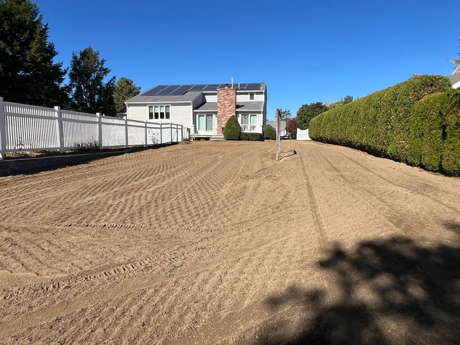 Large backyard with prepared dirt surface, white fence on the left, tall hedges on the right, and a house with solar panels on the roof in the background.