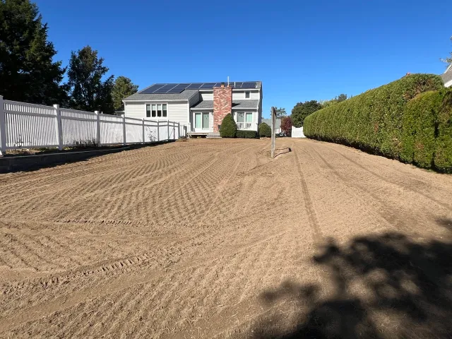 Large backyard with prepared dirt surface, white fence on the left, tall hedges on the right, and a house with solar panels on the roof in the background.